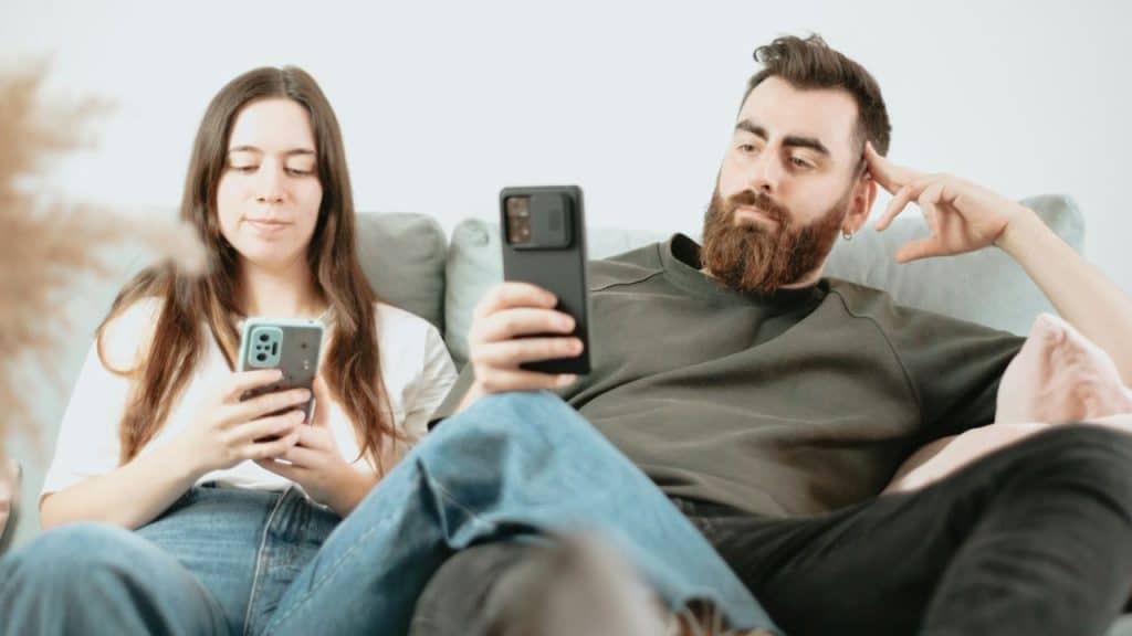 A man and a woman sit on a couch next to each other, both looking at their mobile phones.