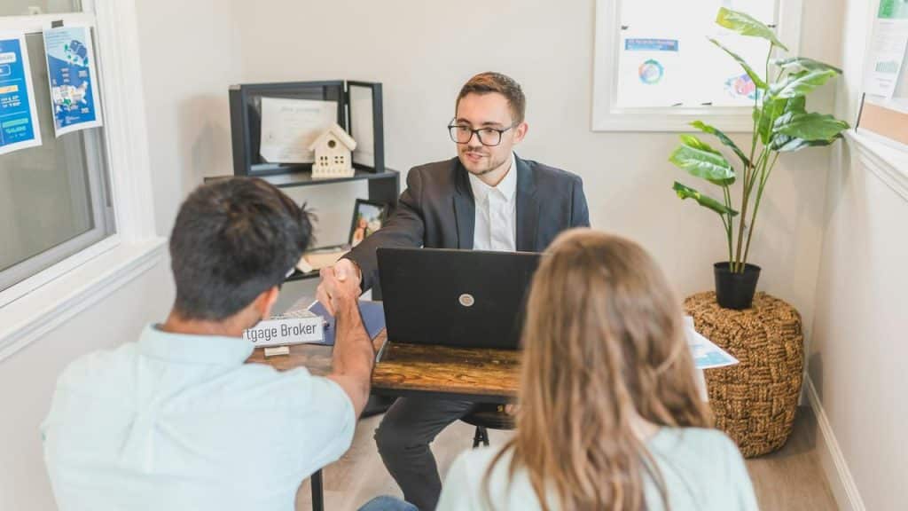 A mortgage broker shakes hands with a couple at a desk.