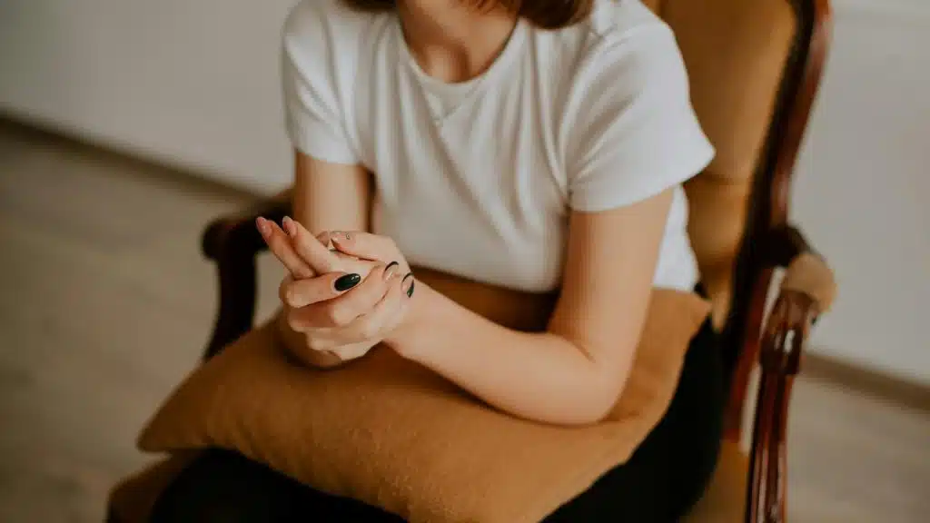 A woman sits on a chair holding her hands together with a pillow on her lap.