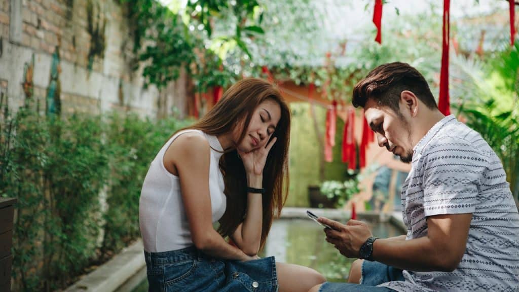 A man is playing a game on his phone while his frustrated wife is sitting with him.