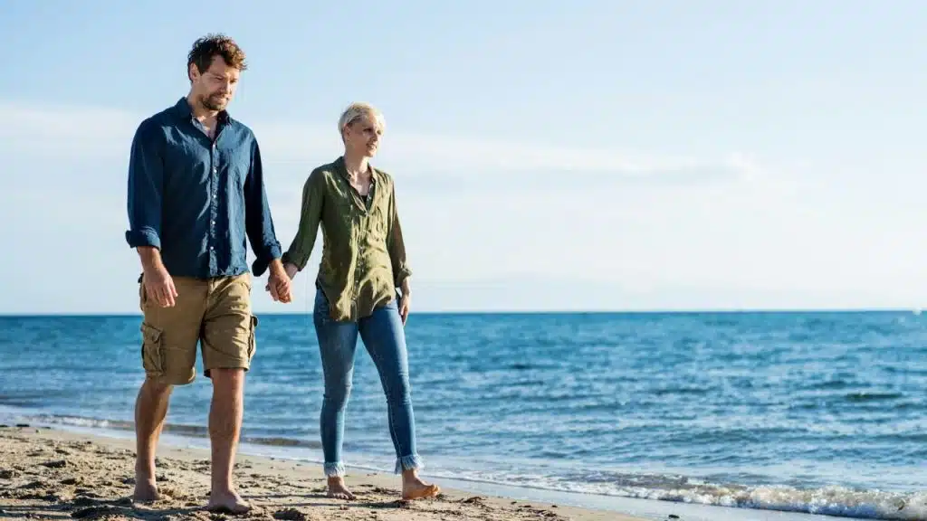Couple holding hands and walking barefoot on a sunny beach near the ocean.