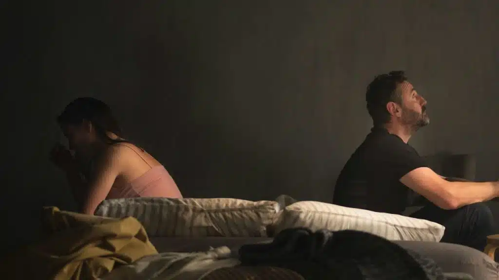 A man and woman sit turned away from each other on a dark bed.