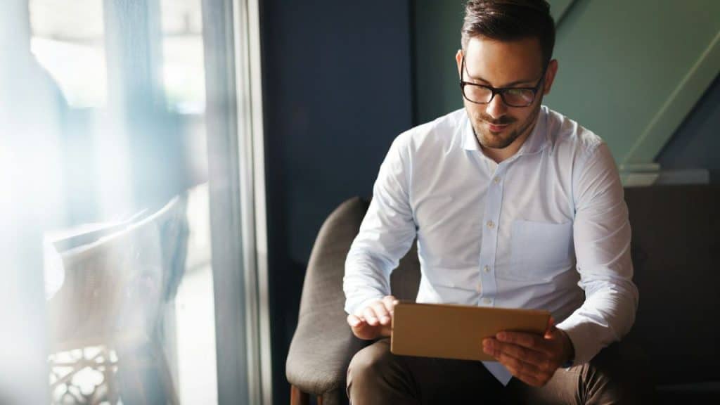 Man in glasses and a white collared shirt looking down at a tablet.