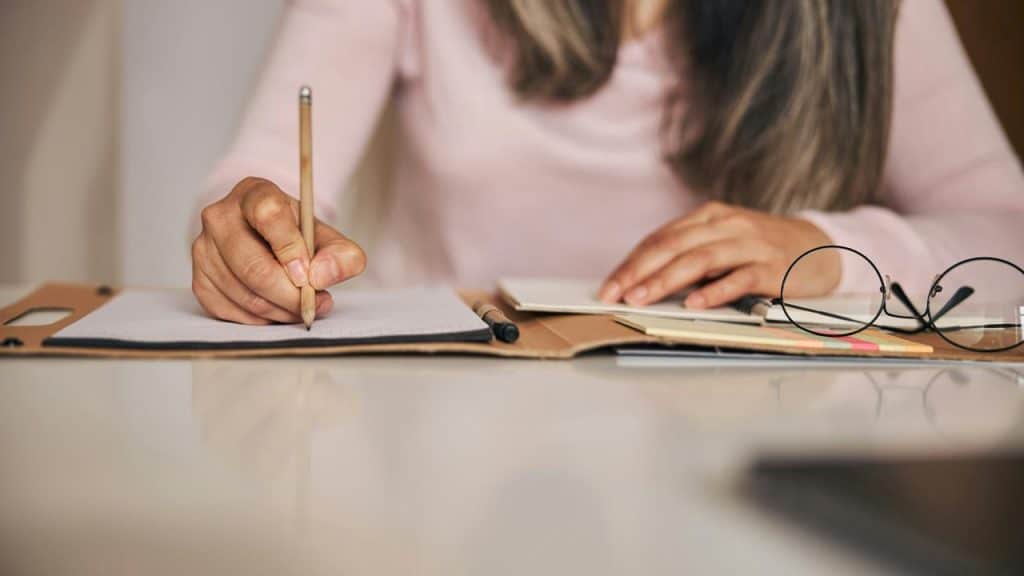 A woman writing in a notebook at a desk with glasses beside her.