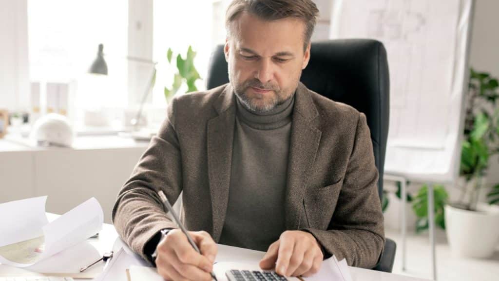 Man in brown suit and turtleneck writing and using a calculator at a desk.