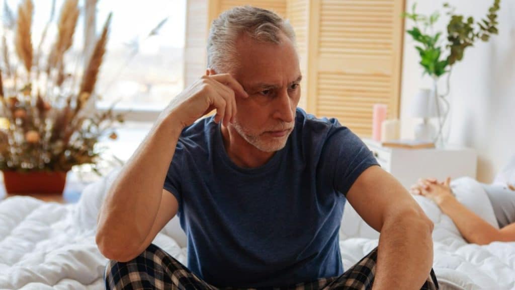 Elderly man with gray hair sits on a bed, looking down, while a woman lies behind him.