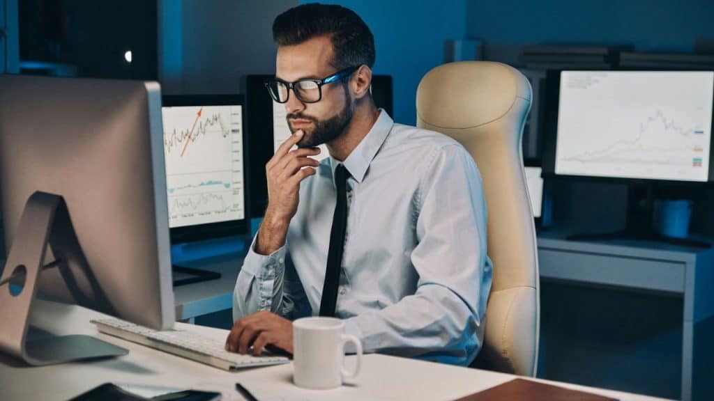 Bearded man in glasses and tie studies a computer screen in a dimly lit office.