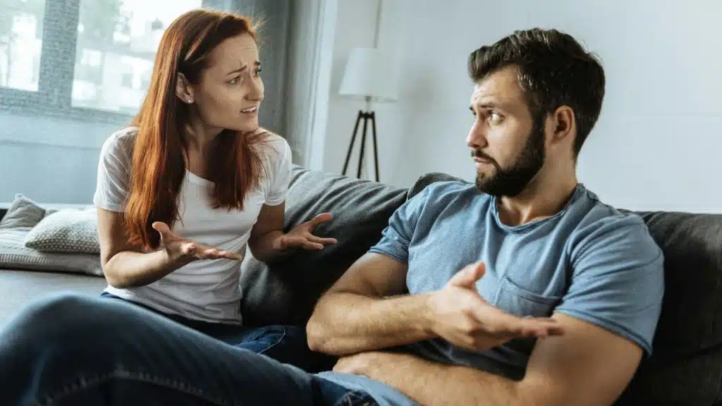 A woman sits on the left, gesturing in frustration at a man on the right who is gesturing defensively.