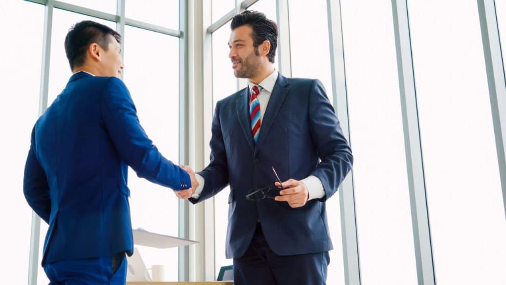 Two businessmen in suits shaking hands in front of a bright window background.
