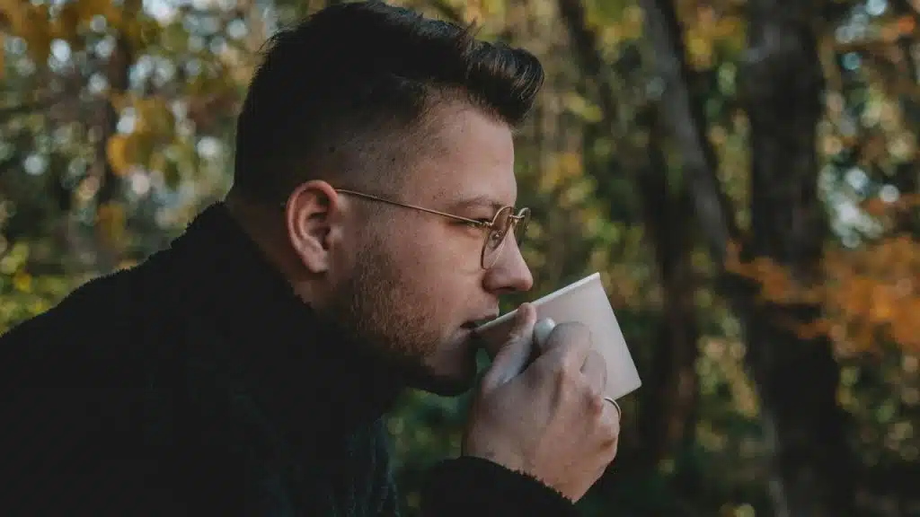 Young man in glasses and dark clothes sips from a cup outdoors with foliage background.