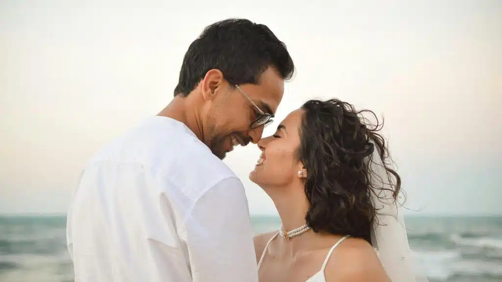 A couple smiling and touching foreheads by the ocean.
