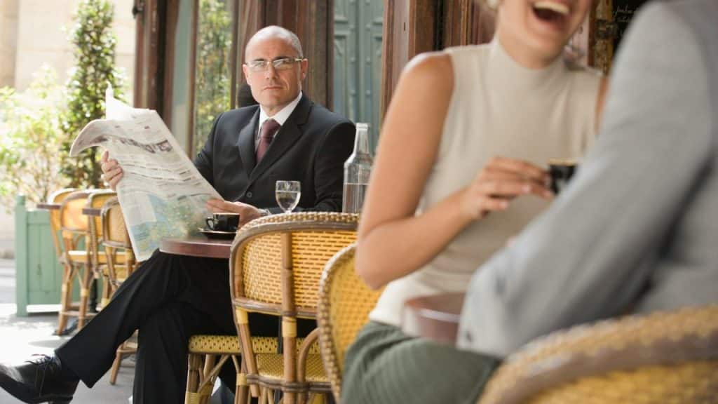 A man in a suit reads a newspaper at a cafe while looking toward the camera.