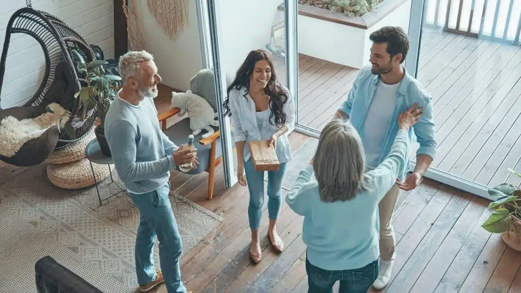 Four adults smile and greet each other in a bright room near a glass door.