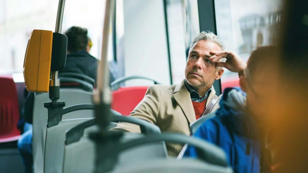A man in a tan coat sits on a bus looking thoughtfully out the window.