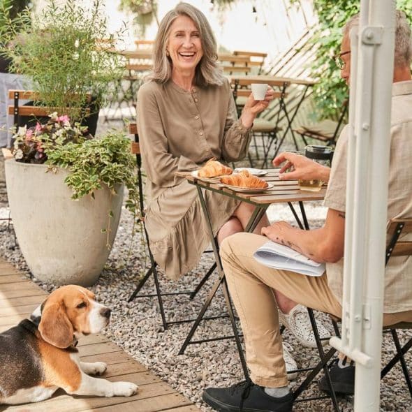 A smiling couple enjoying coffee and croissants at an outdoor café while a dog rests nearby.