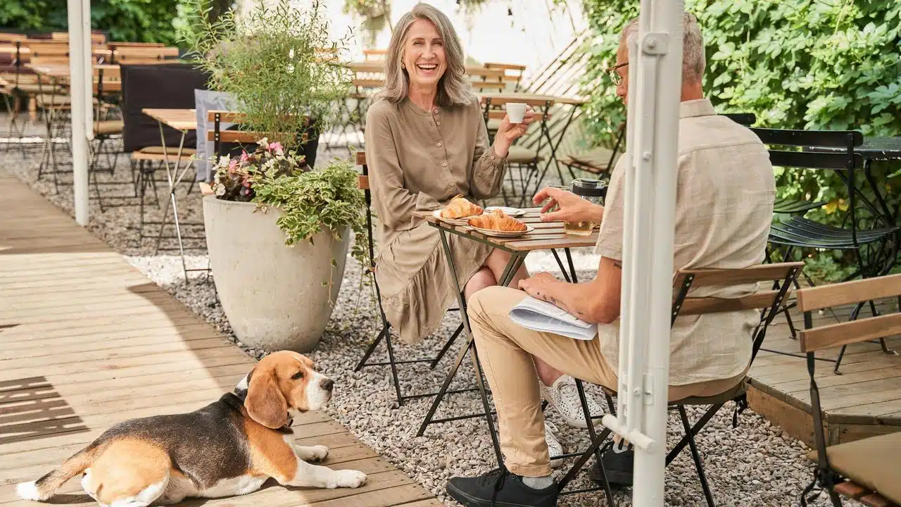 A smiling couple enjoying coffee and croissants at an outdoor café while a dog rests nearby.