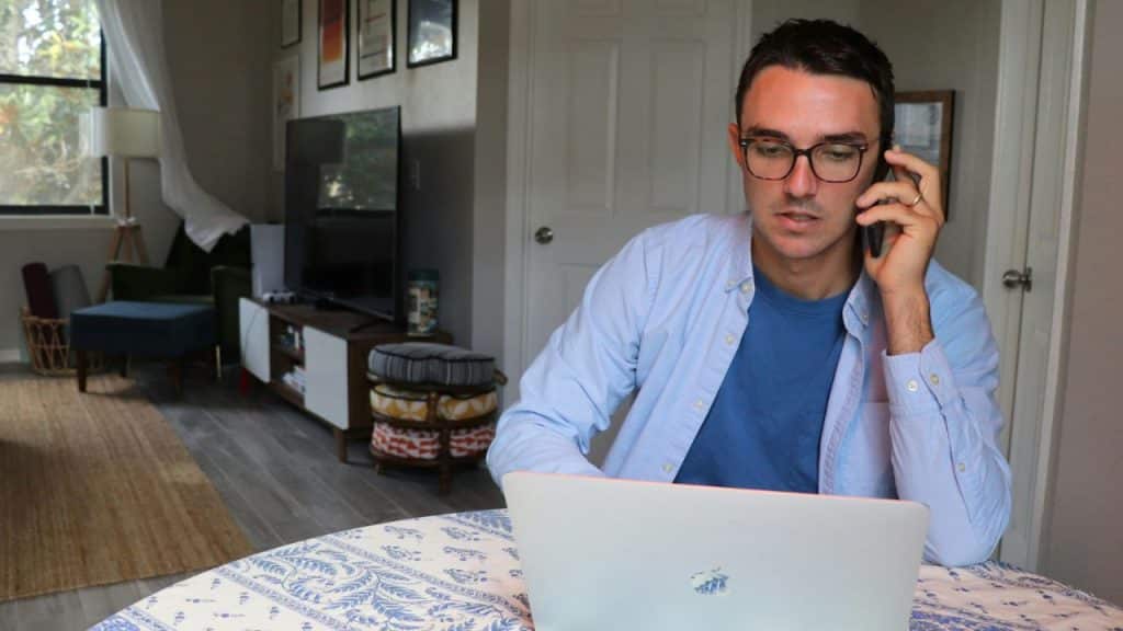 A man wearing glasses and a light blue shirt talks on a phone while using a laptop.
