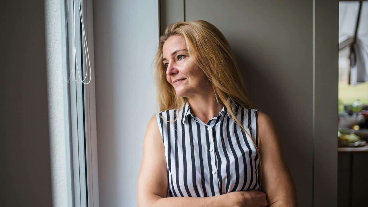 A woman in a striped sleeveless shirt standing by a window and looking outside.