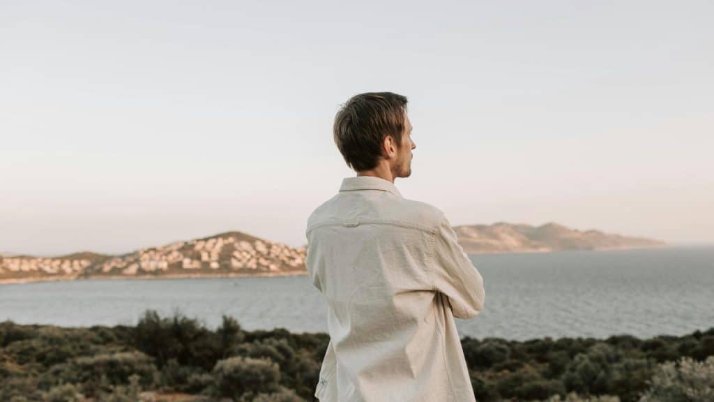 A man standing outdoors, looking out over the sea and distant hills.