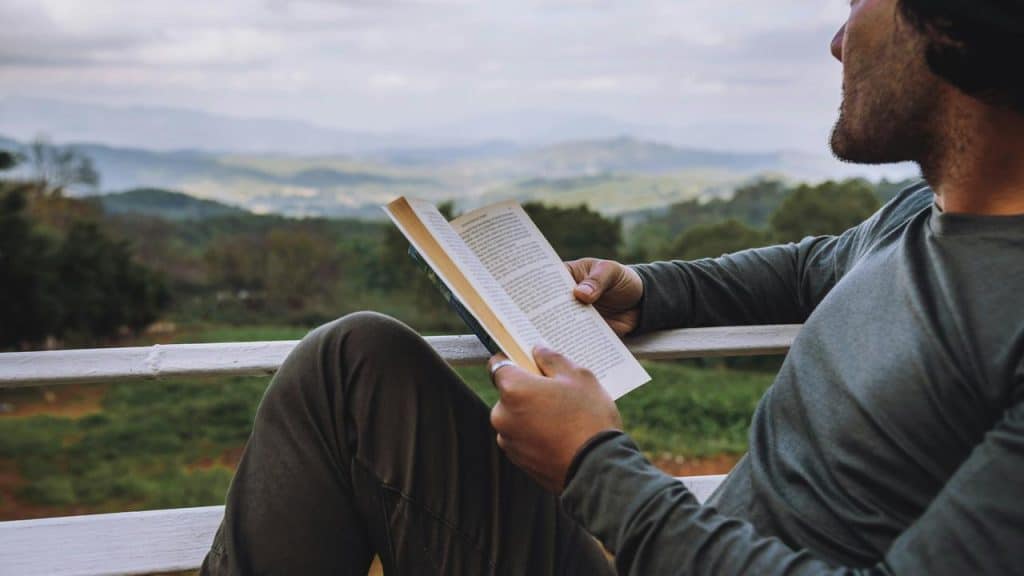 A man sitting outdoors reading a book with a scenic mountain view.