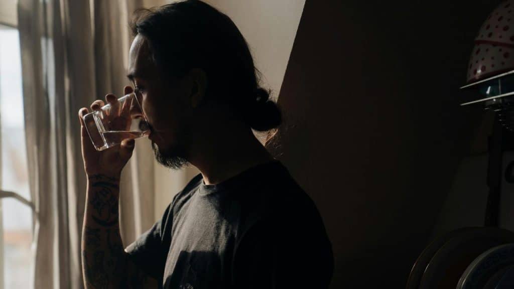 A man standing by a window drinking from a glass in soft light.
