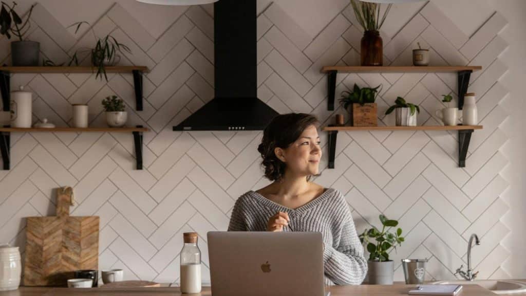 A woman standing in a modern kitchen with a laptop, looking thoughtfully to the side.
