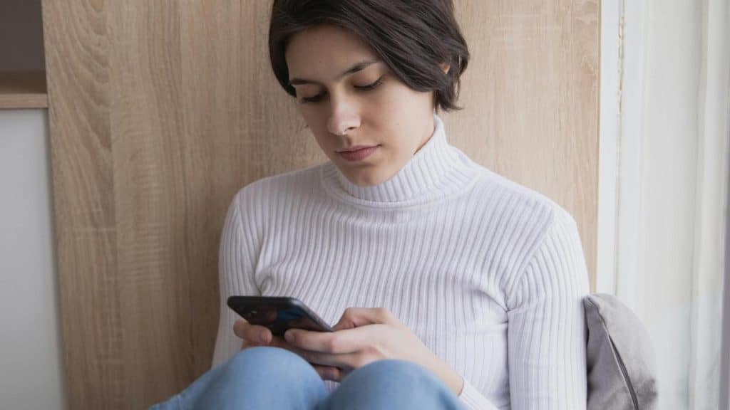 A woman sitting by a wall while looking down at her phone.