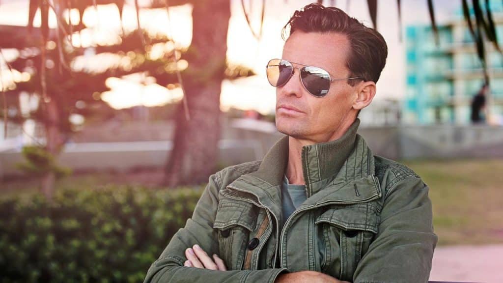 A man wearing aviator sunglasses and a green jacket sits outdoors with his arms crossed.