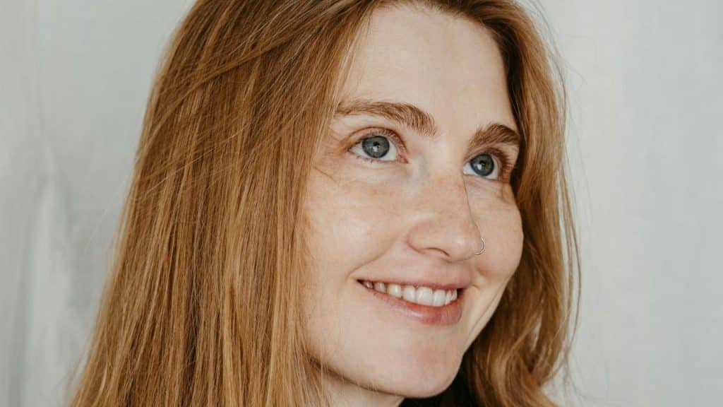 A close-up profile shot of a smiling woman with red hair and light-colored eyes.