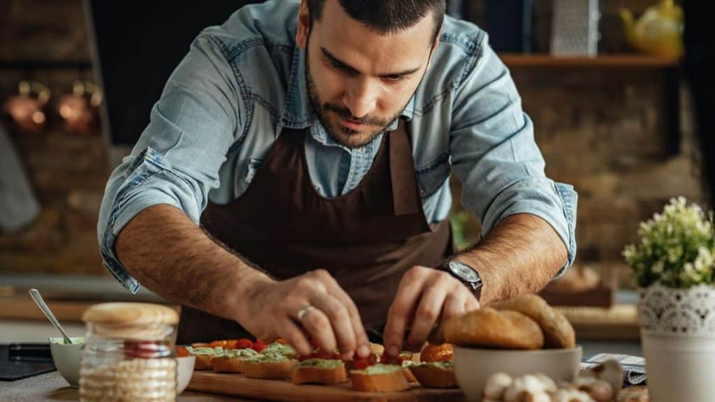 A man in an apron carefully prepares small appetizers on a wooden board.