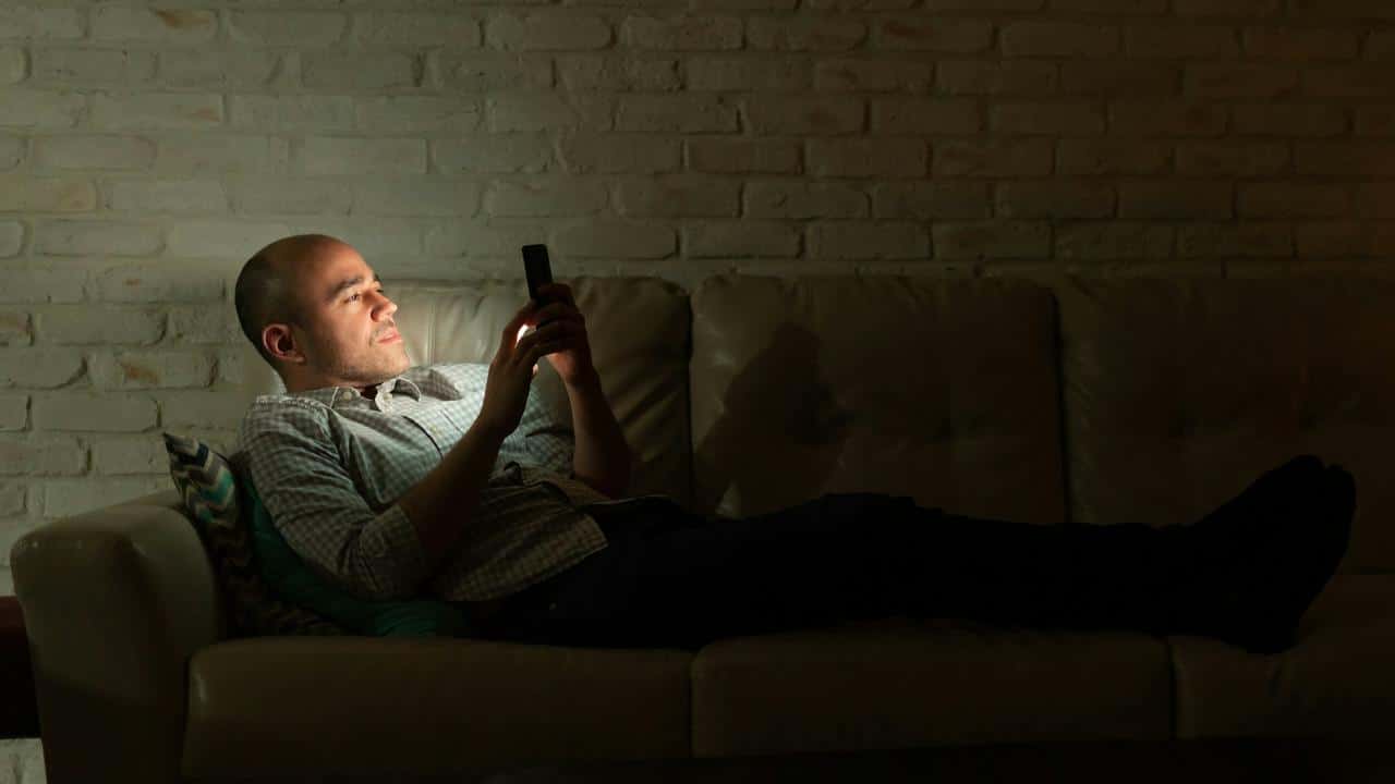 A man lying on a couch in a dark room looking at his phone.