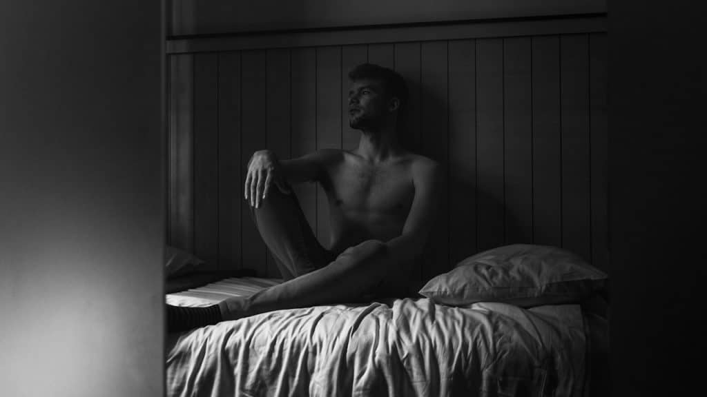 A shirtless man sitting cross-legged on a bed in black and white, looking away thoughtfully.