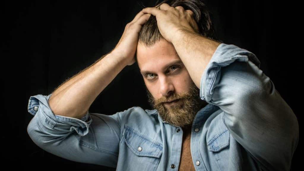 A bearded man in a denim shirt holds his hands up to his dark hair, looking intently forward.