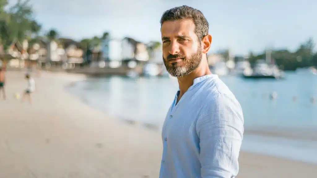 A man with a beard and light blue shirt standing on a beach, looking toward the camera.