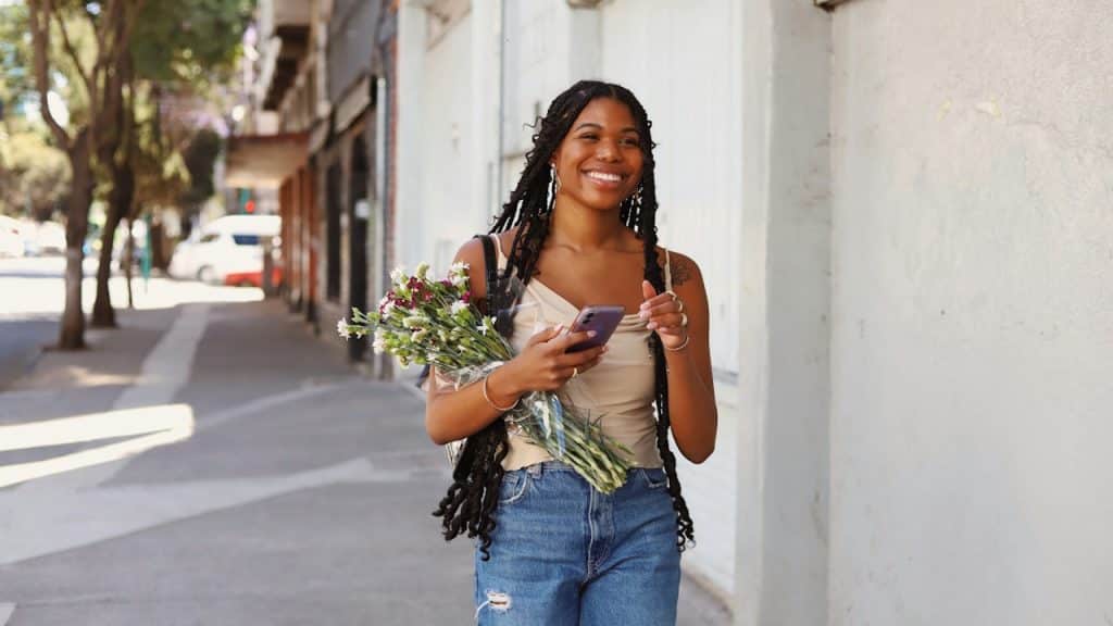 A young woman with long dark braids, wearing a beige tank top and ripped jeans, is walking on a sunny urban sidewalk.
