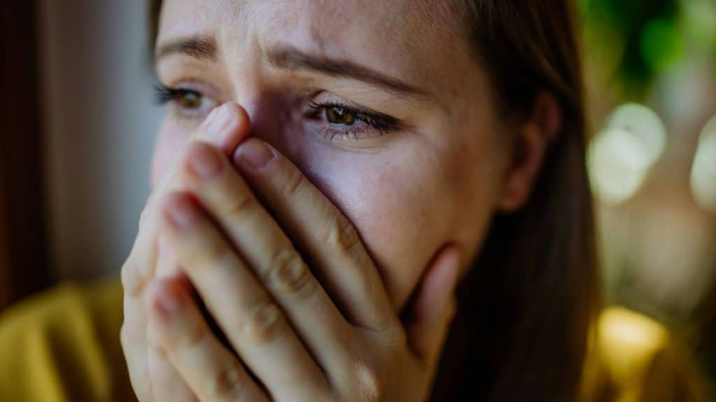 A close-up of a woman's face, her eyes filled with tears, covering her mouth with her hands.
