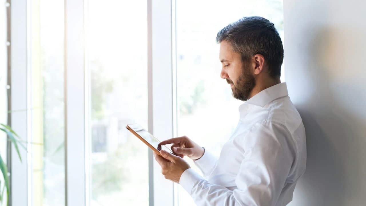 Bearded man in a white shirt using a tablet next to a bright window.