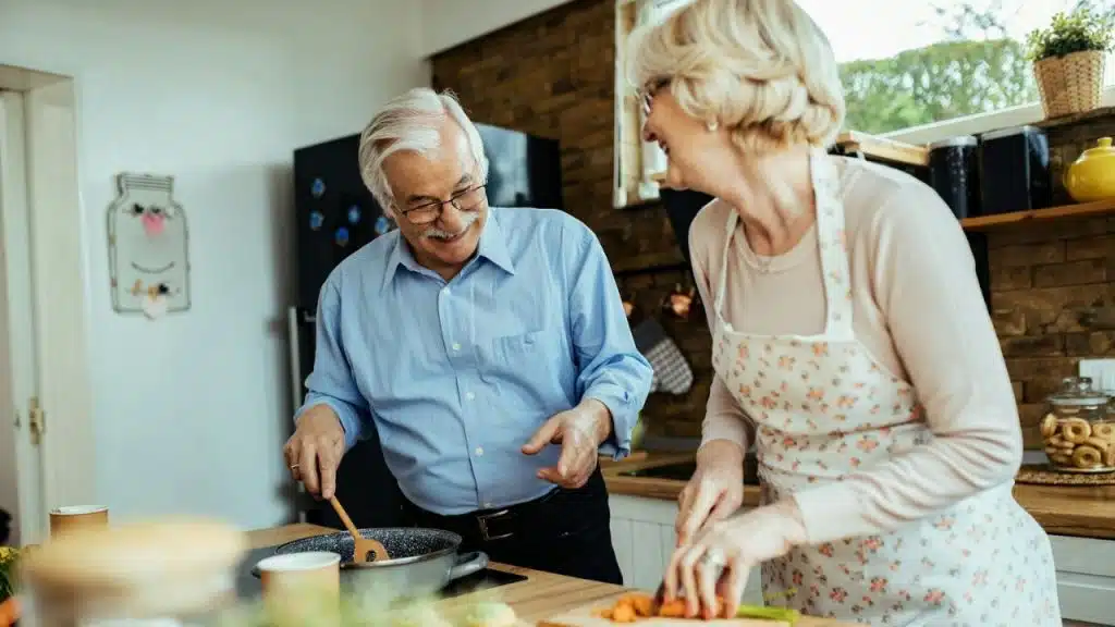 Older smiling couple cooking together in a kitchen with one stirring and one chopping.
