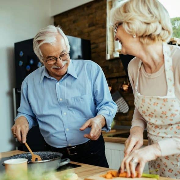 Older smiling couple cooking together in a kitchen with one stirring and one chopping.