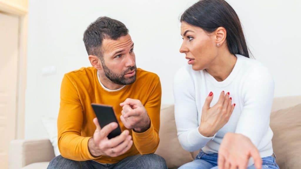 Man holding a smartphone arguing with a woman who is gesturing with her hands.