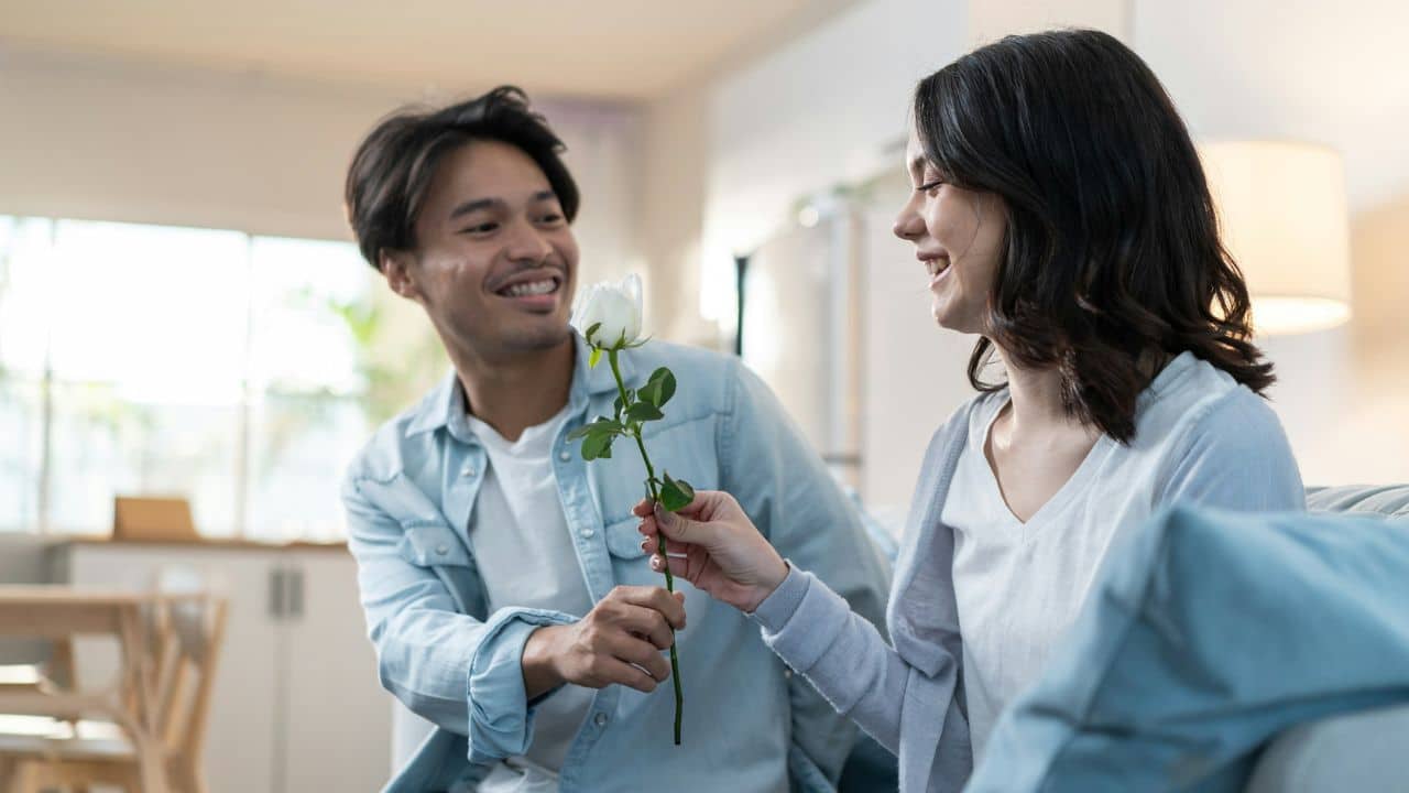 A smiling man hands a single long-stemmed white rose to a smiling woman on a couch.