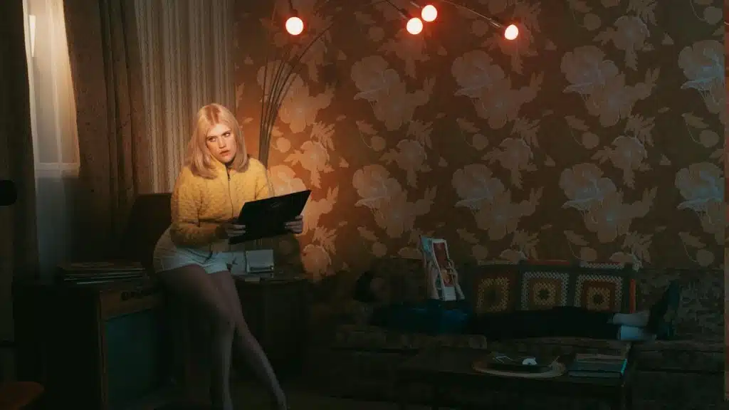 A woman in a yellow long-sleeve shirt holding a tablet while sitting on a chair in a dark room.