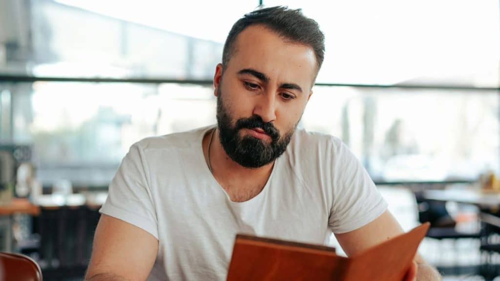 Young man with a beard in a white shirt looking intently at a menu.