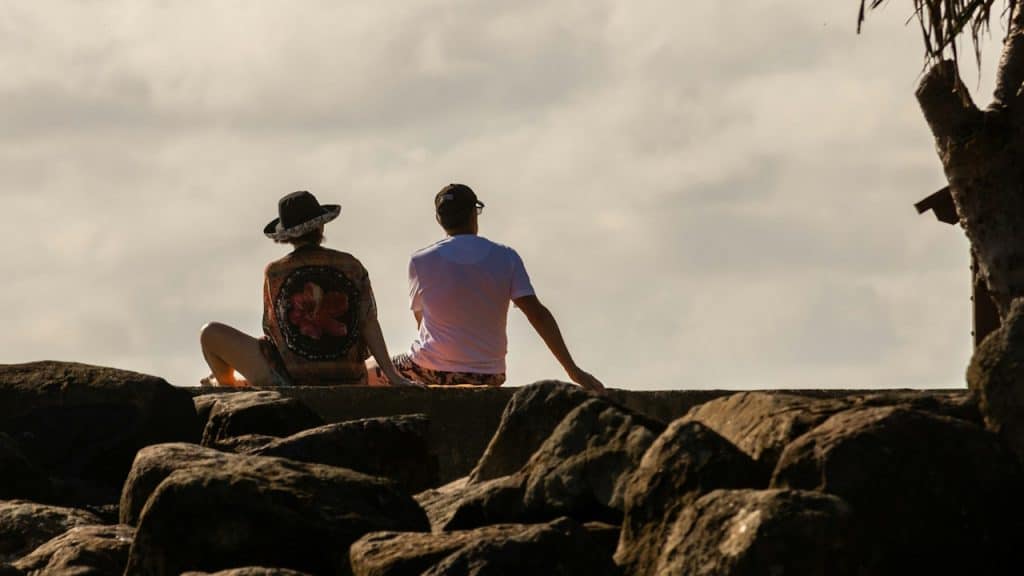 Two people sit on large rocks near the shore, facing away from the camera as they look out toward a cloudy sky.