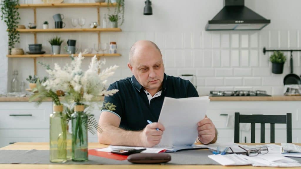 Older man with glasses studying financial documents at a kitchen table.