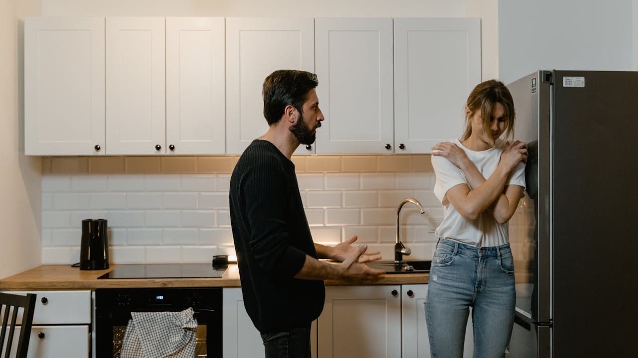 A man is arguing with a woman who is leaning against a refrigerator with her arms crossed over her chest.
