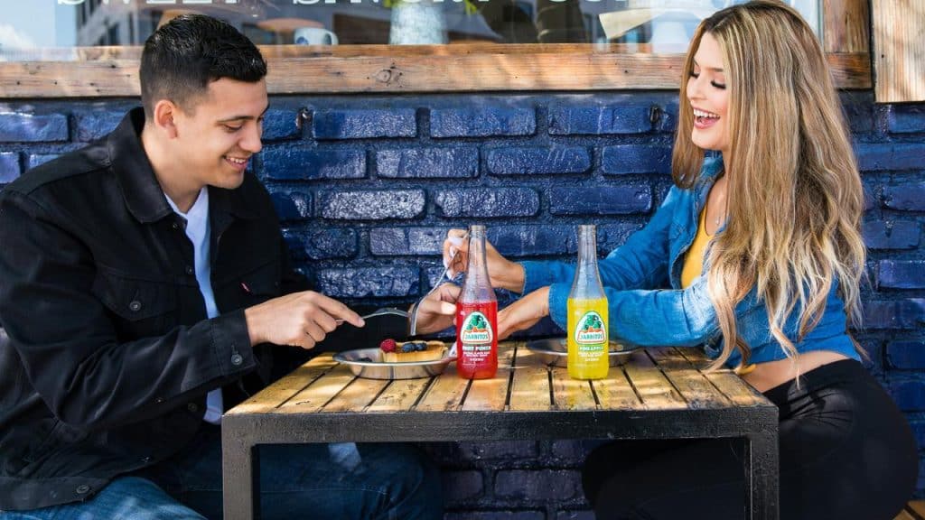 A man and woman smiling while sharing food and drinks at an outdoor table.