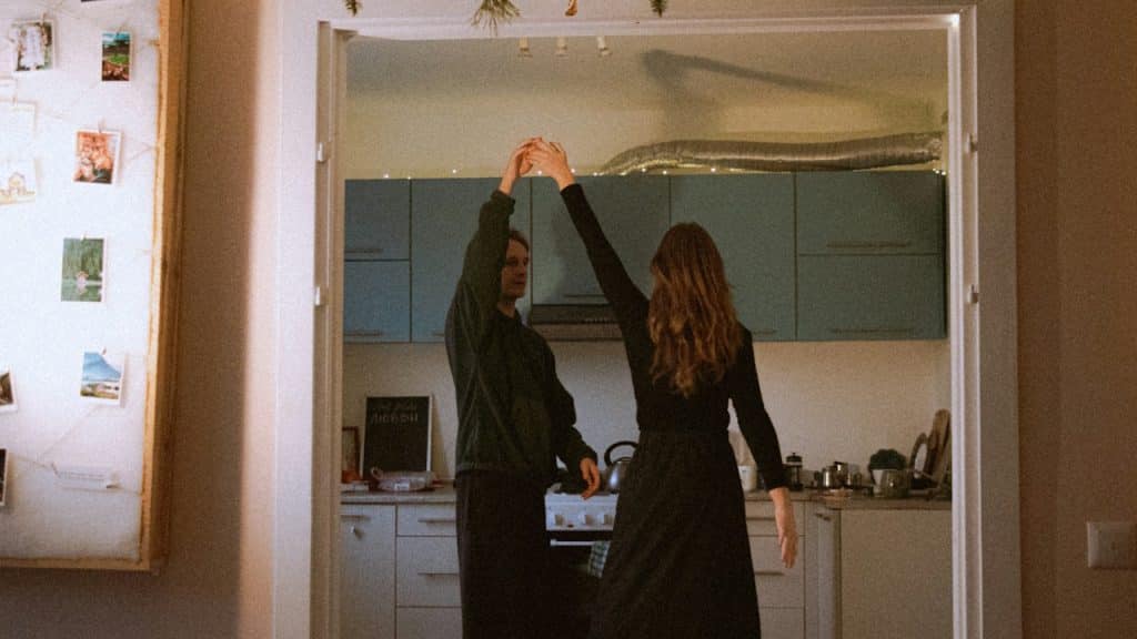 A couple is dancing in a doorway, holding hands over their heads, with a kitchen visible in the background.