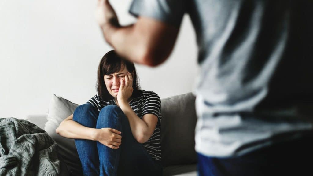 A crying woman covers her face while a blurred man is aggressively gesturing in the foreground.
