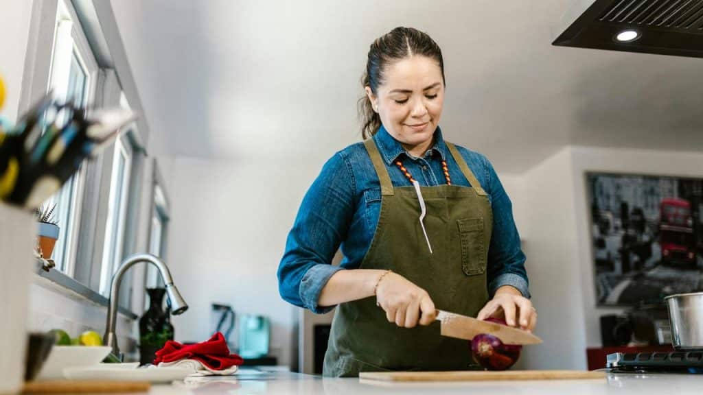 A woman chopping a vegetable in a bright kitchen while wearing an apron.